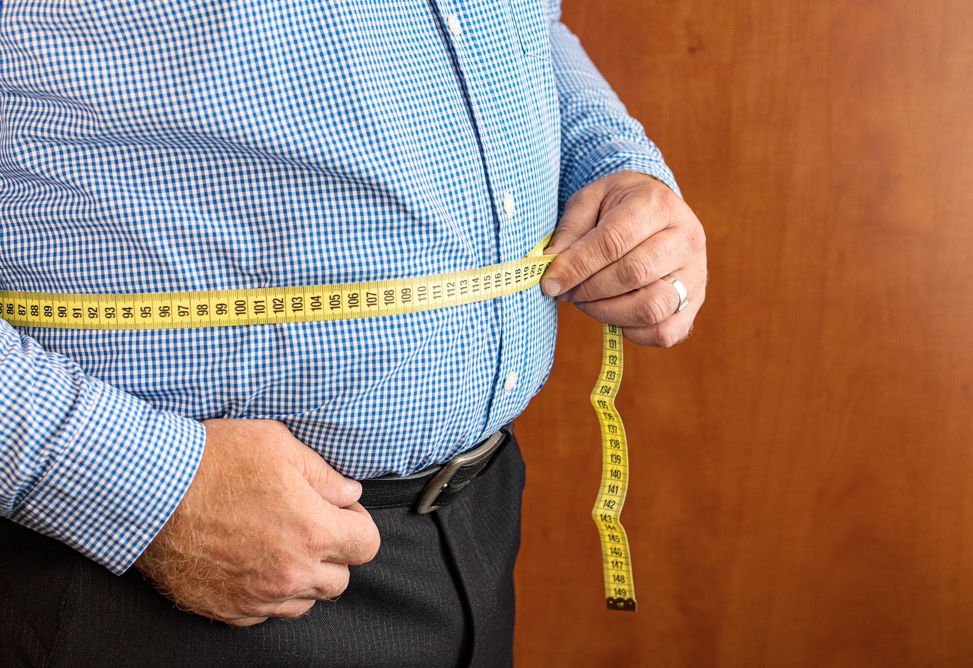 Mann misst seinen Bauchumfang mit einem gelben Maßband, trägt ein kariertes Hemd und dunkle Hose.