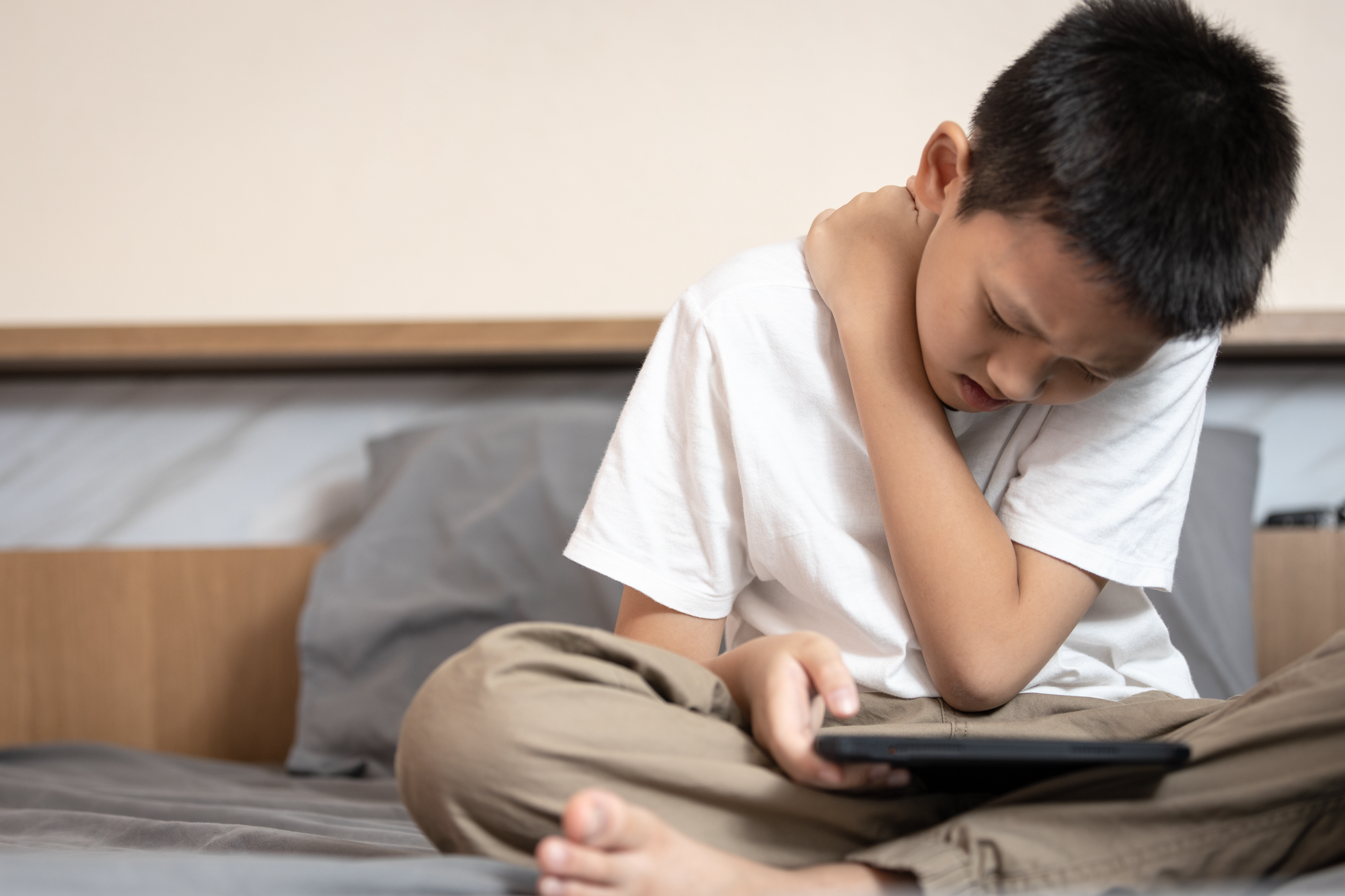 rueckenfit.png Ein Junge sitzt auf einem Bett, hält ein Tablet und fasst sich schmerzverzerrt an den Hals.