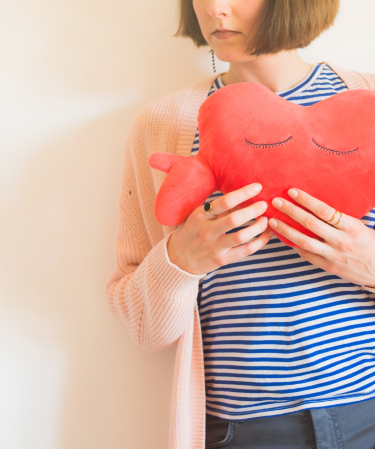 Eine Person hält ein rotes herzförmiges Kissen mit geschlossenen Augen, gekleidet in gestreiftes Oberteil und eine helle Strickjacke.