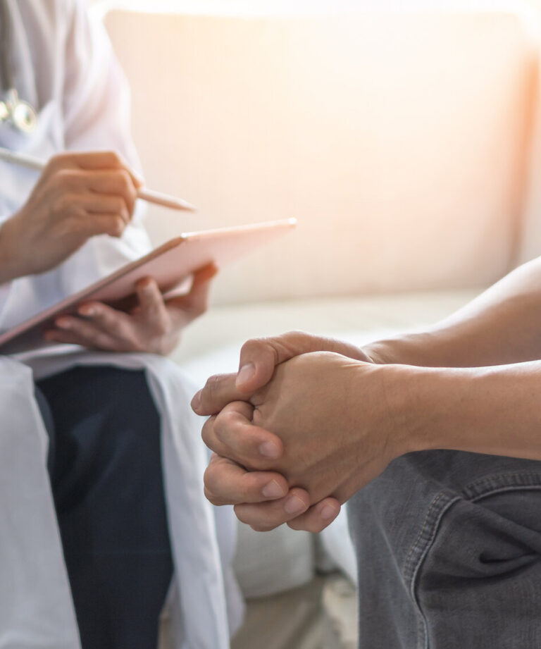 Arzt in weißem Kittel notiert auf einem Tablet, während ein Patient mit zusammengefalteten Händen sitzt.