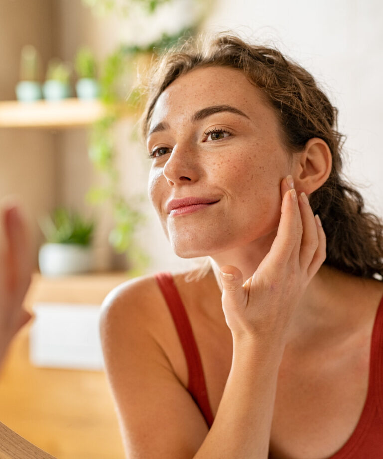 Frau mit lockigem Haar, die im Spiegel ihre Haut berührt, sanfte Beleuchtung und Pflanzen im Hintergrund.