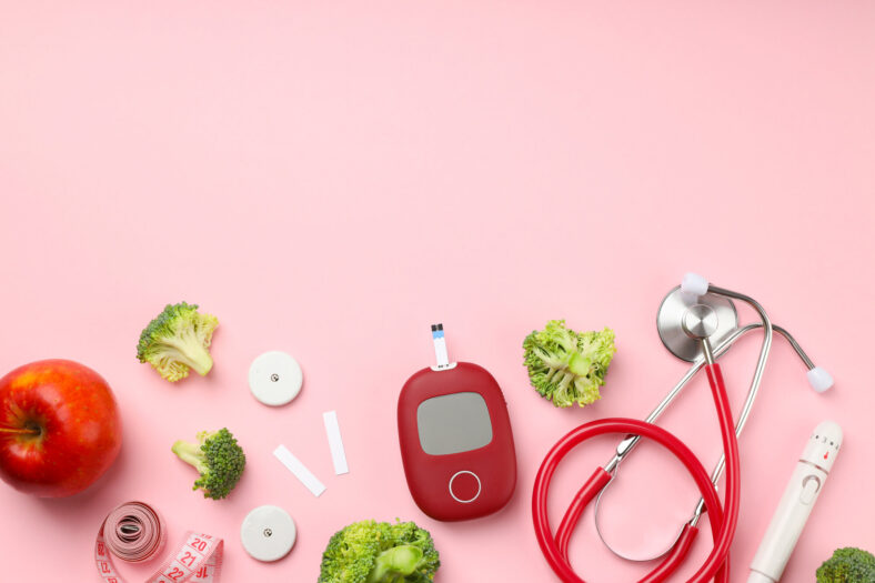 Gesunde Lebensmittel und medizinische Geräte auf rosa Hintergrund, darunter Apfel, Brokkoli, Blutzuckermessgerät und Stethoskop.
