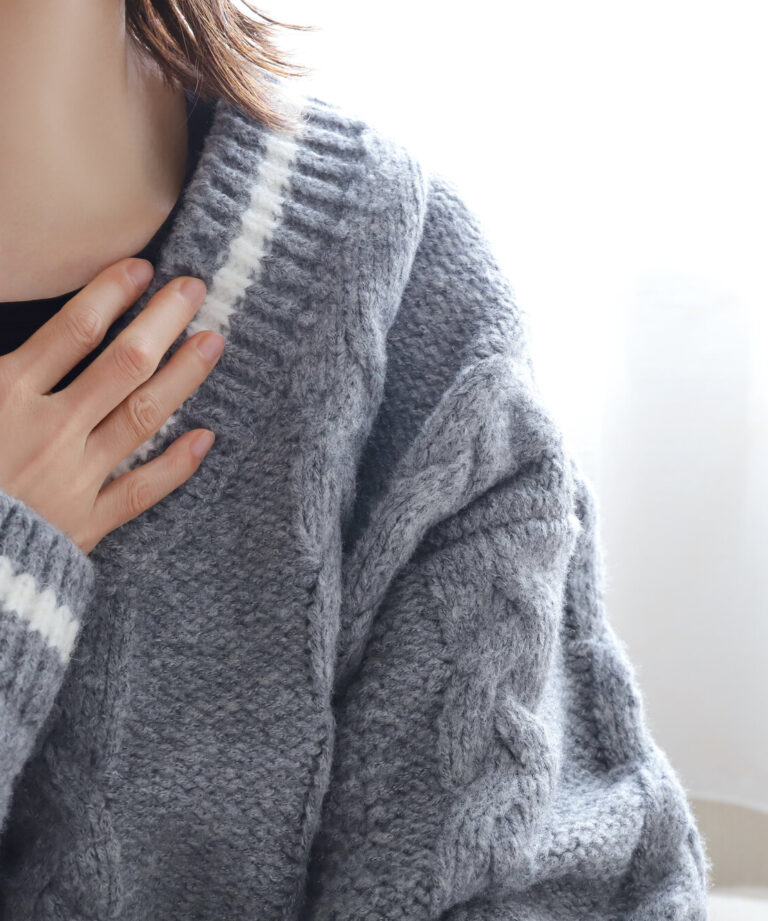 Eine Person in einem grauen, gestrickten Pullover mit weißen Streifen zeigt eine nachdenkliche Geste.