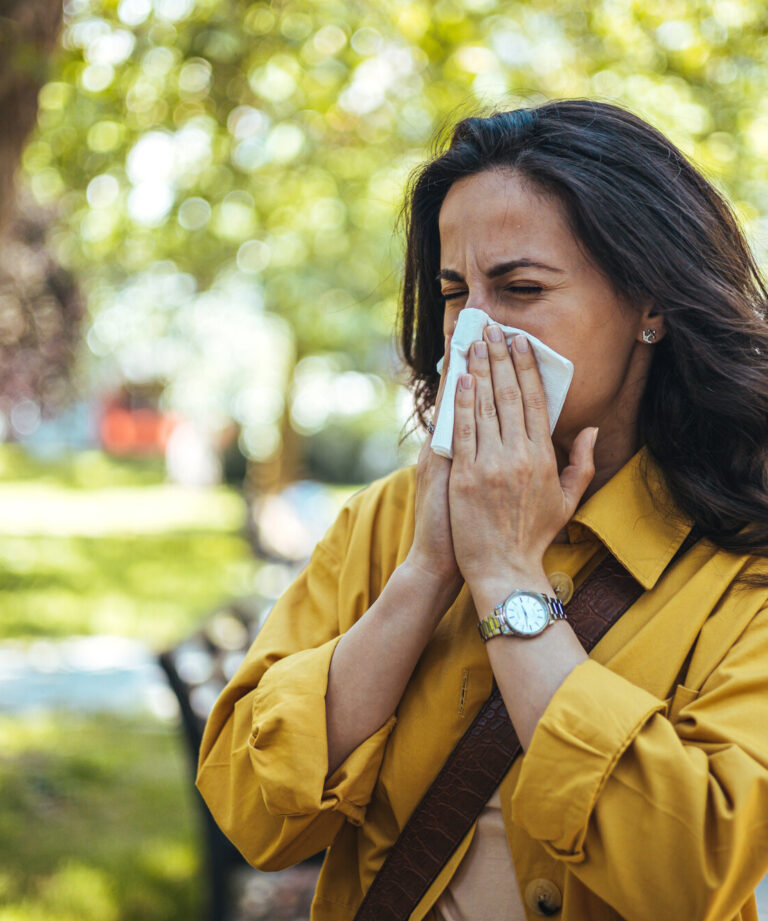 Frau in einem gelben Hemd, die sich mit einem Taschentuch die Nase putzt, umgeben von grüner Parklandschaft.