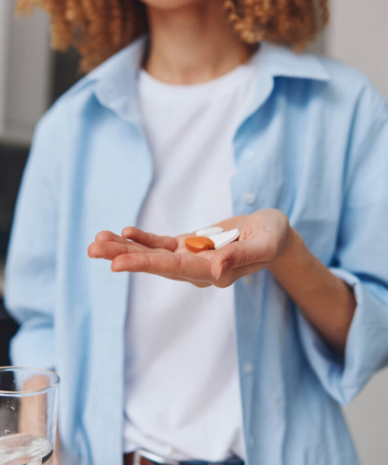 Eine Person hält zwei Kapseln in einer Hand, während in der anderen ein Glas Wasser steht, in einer modernen Küche.