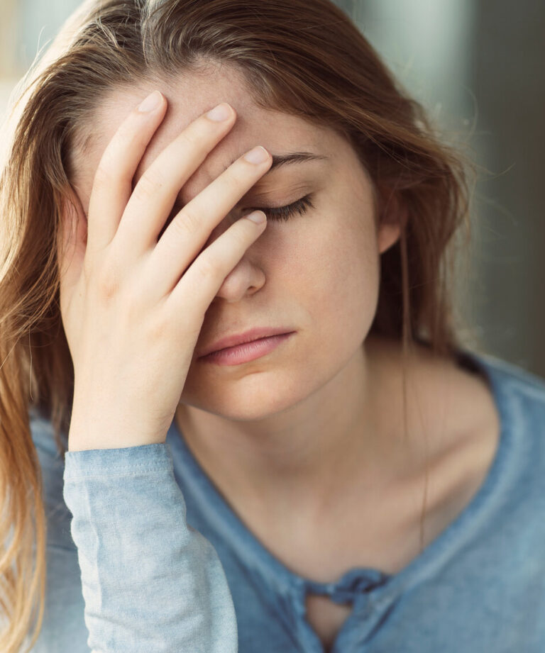 Junge Frau mit langen Haaren und blauem Oberteil, die frustriert ihre Hand an die Stirn legt, im Innenraum sitzend.
